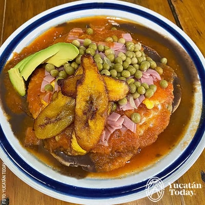 Presentation of Huevos Motuleños. This traditional Yucatecan dish consists of a tortilla with black beans and a fried egg topped with tomato sauce, ham, cheese, peas, and plantains.